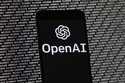 The OpenAI logo appears on a mobile phone in front of a computer screen with random binary data, Ma…