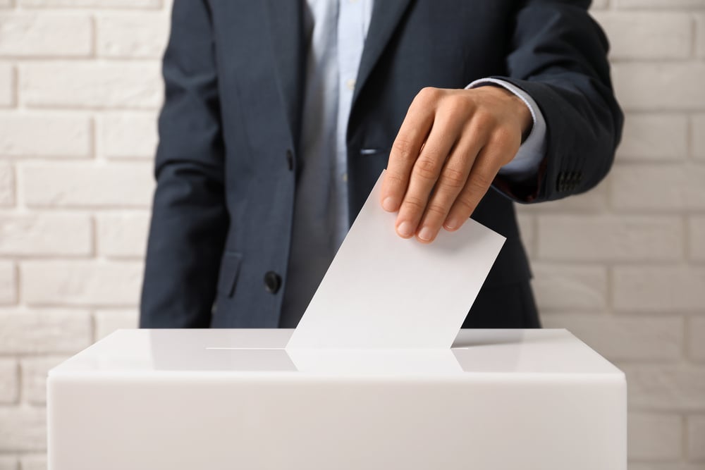 Man putting his vote into ballot box against brick wall: learn more about election stocks