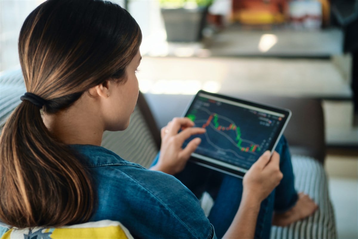 Young adult woman lying down on sofa in living room at home, trading online with tablet computer connected to stock markets. Over the shoulder view