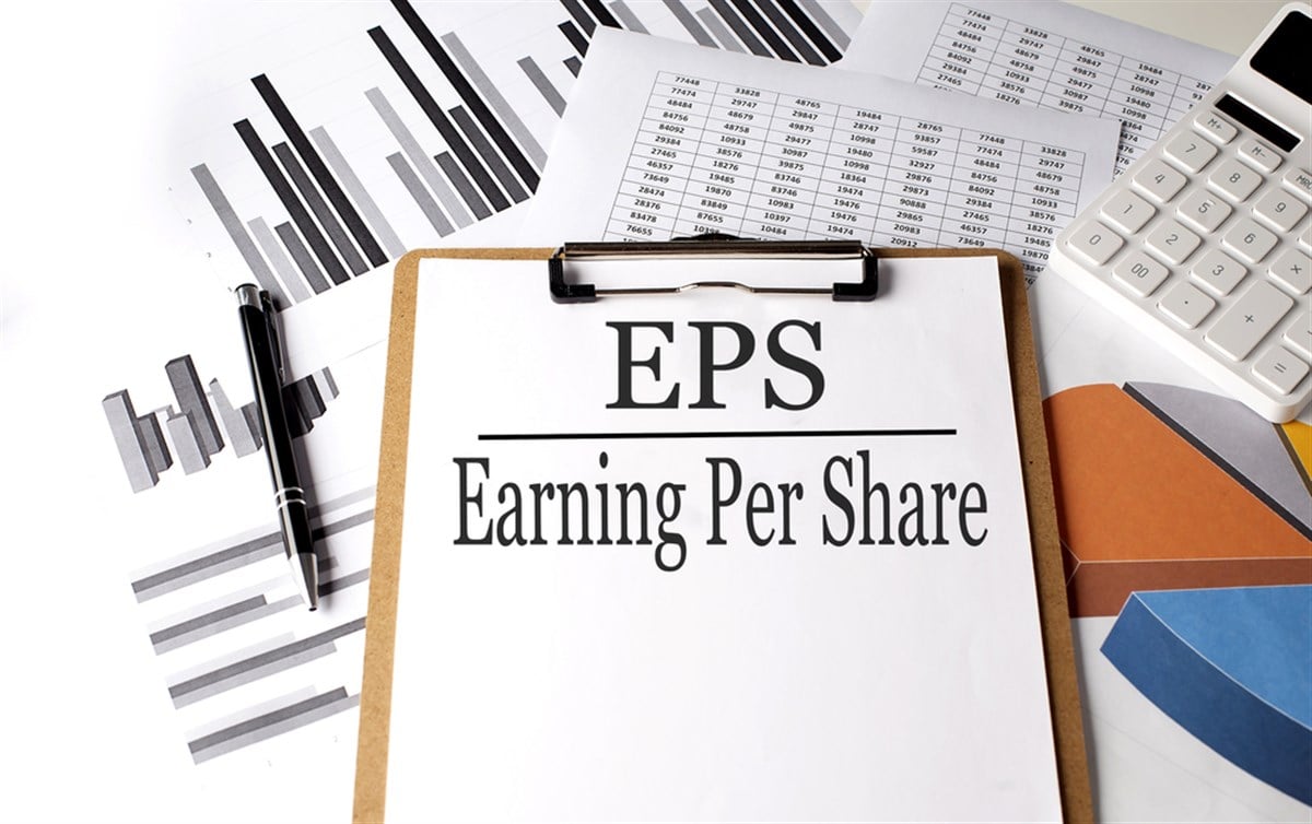 Photo of a clipboard with "EPS Earnings Per Share" on a sheet of paper; charts and a calculator behind it.