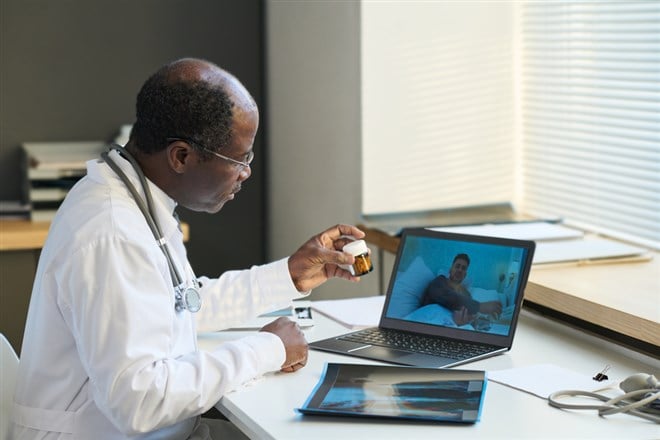 Mature African American general practitioner recommending pills to online sick male patient on screen of laptop during consultation