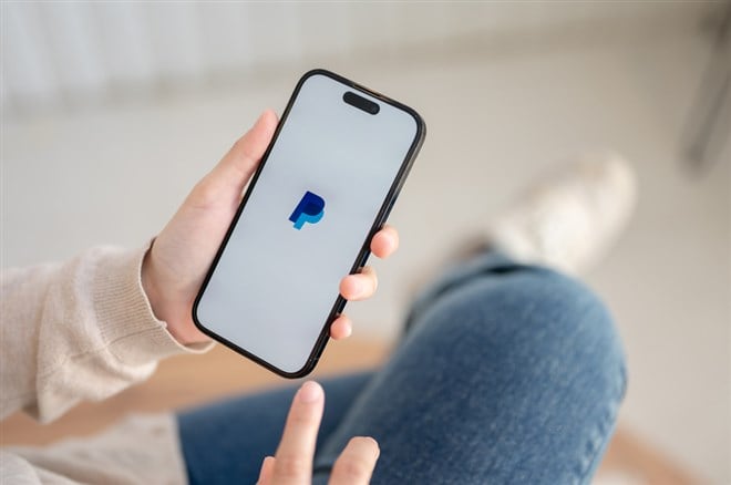 A woman sitting indoors and using a PayPal application through her Iphone14. A PayPal logo on an Iphone14 screen.