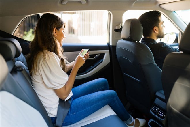 Side view of an attractive latin woman sitting on the car back seat and using a ride share app on her smartphone 