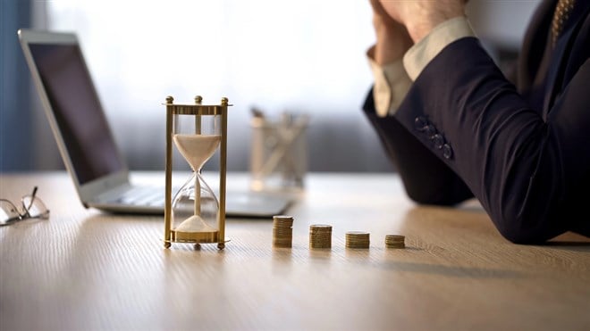 Photo of a man in a suit at a laptop with a sandglass and stacks of coins symbolizing how patience pays off with a buy-and-hold investment strategy.