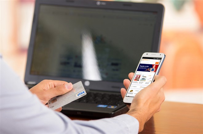 Closeup of young mans hands holding smartphone up with Alibaba website, Visa card in other hand with laptop computer