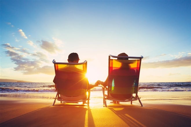 Photo of a retired couple sitting in chairs holding hands on a beach during a sunset