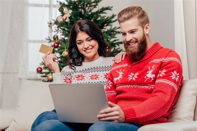 Photo of a couple in Christmas sweaters in front of a Christmas tree. Woman has credit card in hand, man has laptop on lap.