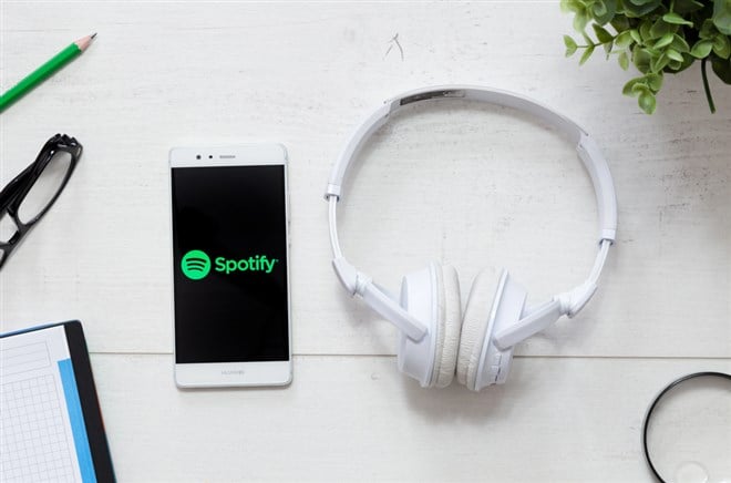 Photo of a smartphone connected to white headphones using the Spotify app on a black background. Green light. Entertainment apps. Streaming.