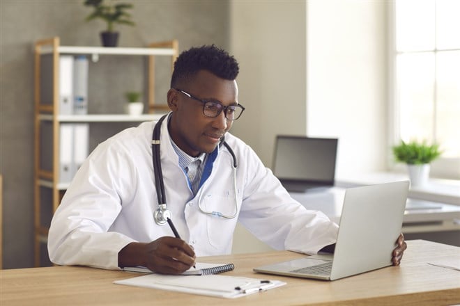 Doctor medical professional looking at laptop screen and writing notes on paper - stock image