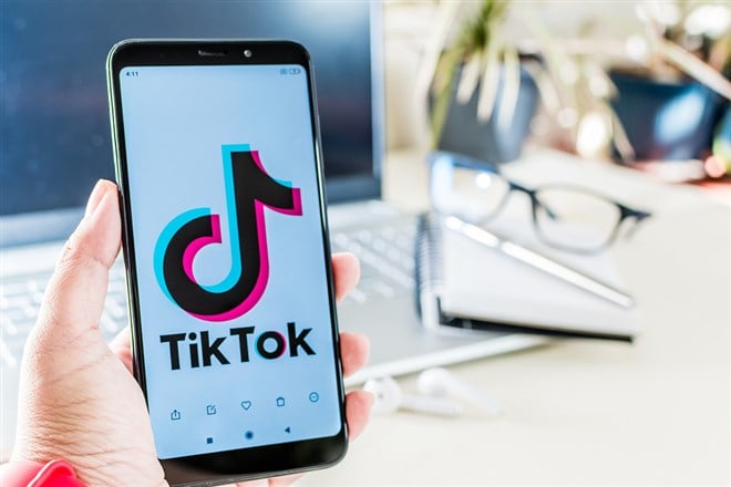 Photo of someone holding a phone with the tiktok logo next to a laptop computer, reading glasses, a notebook, and pen