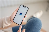 A woman sitting indoors and using a PayPal application through her Iphone14. A PayPal logo on an Iphone14 screen.