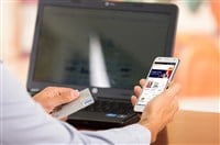 Closeup of young mans hands holding smartphone up with Alibaba website, Visa card in other hand with laptop computer