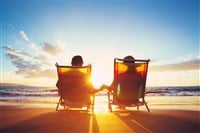 Photo of a retired couple sitting in chairs holding hands on a beach during a sunset