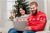 Photo of a couple in Christmas sweaters in front of a Christmas tree. Woman has credit card in hand, man has laptop on lap.