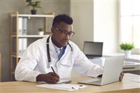 Doctor medical professional looking at laptop screen and writing notes on paper - stock image