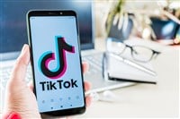 Photo of someone holding a phone with the tiktok logo next to a laptop computer, reading glasses, a notebook, and pen